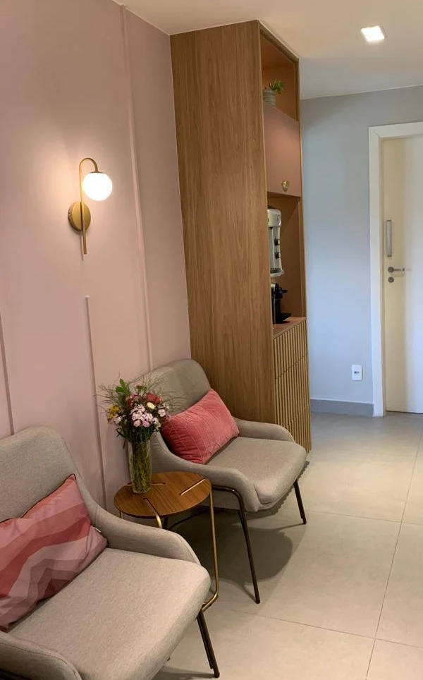 Sala de espera com duas poltronas, almofadas rosadas, uma mesinha com flores e uma arandela dourada na parede rosa.