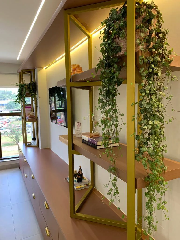 Estante de metal dourado com prateleiras terracota, plantas pendentes e, ao fundo, a janela para o gramado.