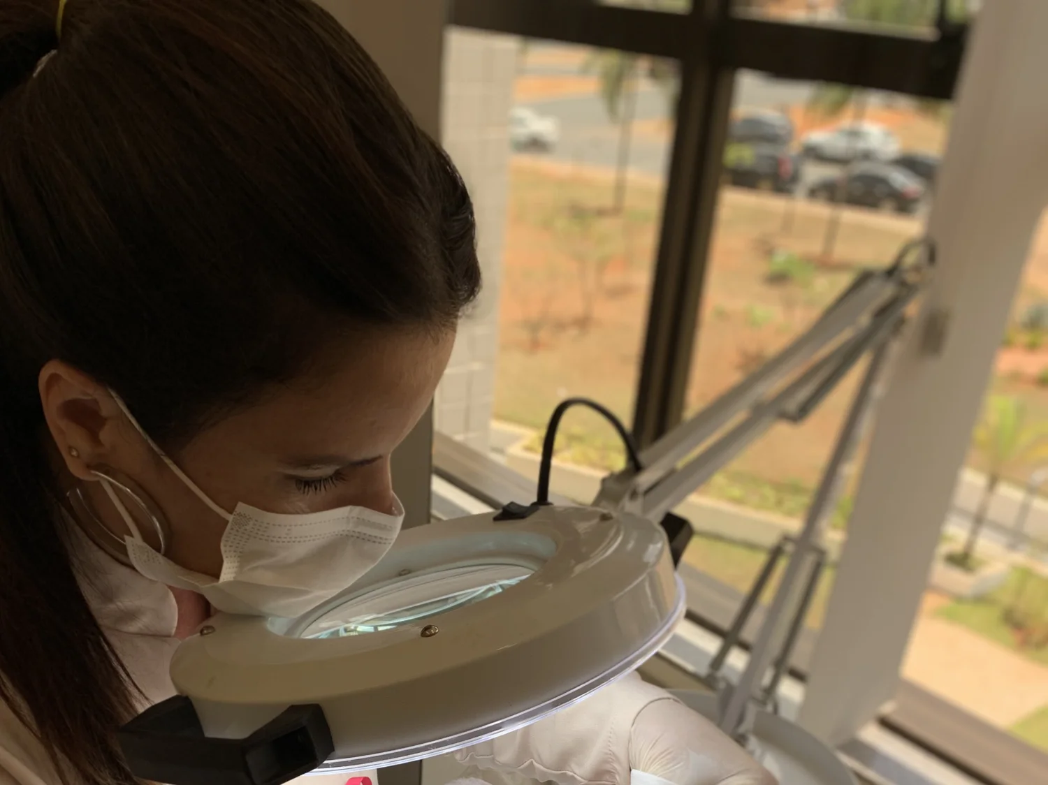 De perto, a profissional de máscara olha pela lupa de luz durante um atendimento facial.