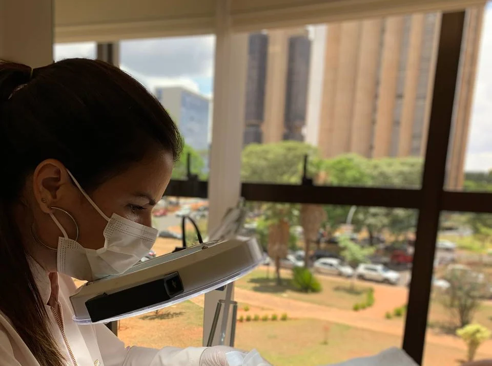 Profissional do Instituto, de máscara, trabalha sob a lupa de luz; pela janela, o verde e os prédios de Brasília.
