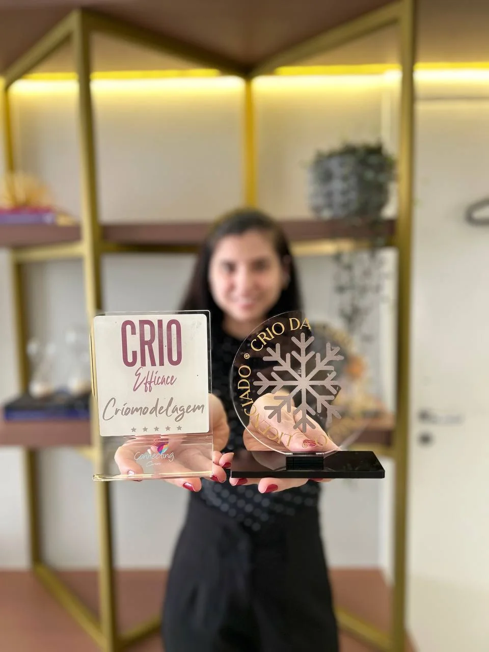 Patrícia, ao fundo e fora de foco, segura à frente dois troféus de criomodelagem.